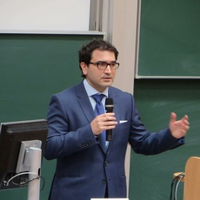 Bild von Prof. Dr. Vassil Tcherveniachki. Er trägt einen blauen Anzug und hält ein Mikro in der Hand. Er hat dunkle kurze Haare und eine Brille.