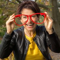 Bild von Romy Einhorn. Sie hält eine rote große Brille in der Hand und lacht. Sie trägt ein gelbes Shirt und eine dunkle Lederjacke.